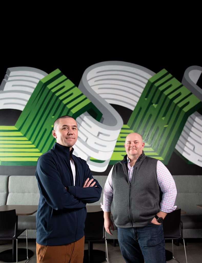VizyPay CEO Austin Mac Nab, left, and Frank Pagano, executive sales director, revel in a high-energy working environment. Behind them the mural sports one of their company mottos: Work hard, play hard.